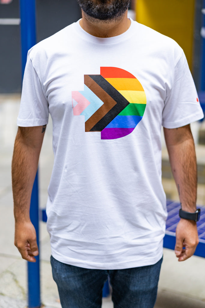 Close-up front view of the Progress Pride Tee in white featuring a large, centrally printed RMIT pixel logo in rainbow colours. The model is standing outdoors in natural light. Available at RMIT Store.
